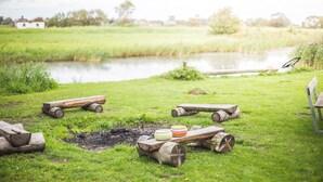 Een rustieke buitenomgeving met een houten vuurkuil, houtblokken en een bank naast een vijver.