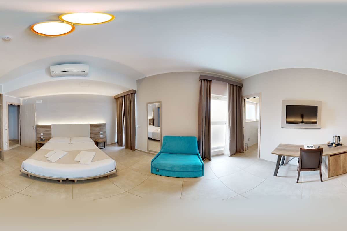 Une chambre d’hôtel moderne avec un grand lit, un fauteuil bleu, un bureau avec une chaise et un grand miroir.
