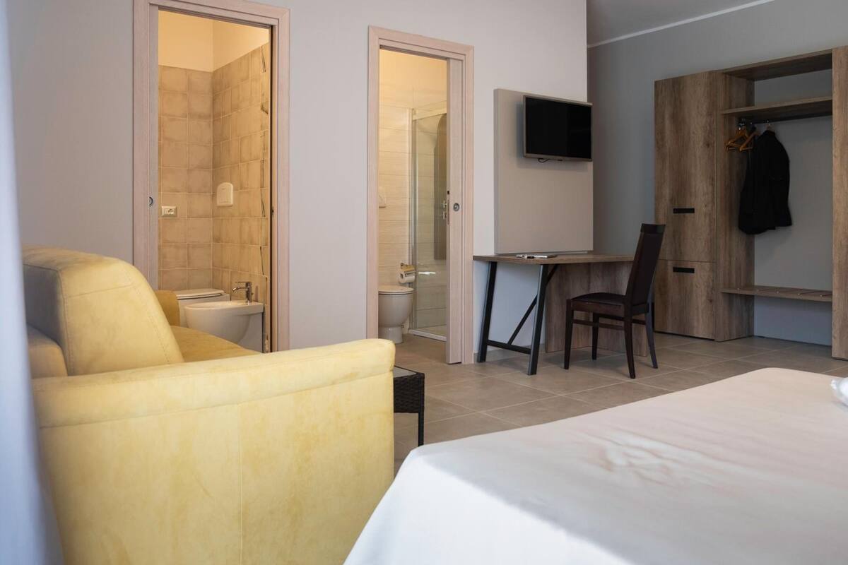 Une chambre d’hôtel avec un lit, un fauteuil jaune, un bureau avec une chaise, un téléviseur fixé au mur et une armoire en bois.