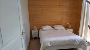 Un dormitorio con cabecera de madera, cama blanca con colcha estampada, dos mesitas de noche con lámparas y una puerta blanca.