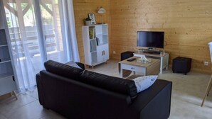 Un salón con un sofá negro, un televisor sobre un mueble bajo, un armario blanco y un ventanal que da al exterior.