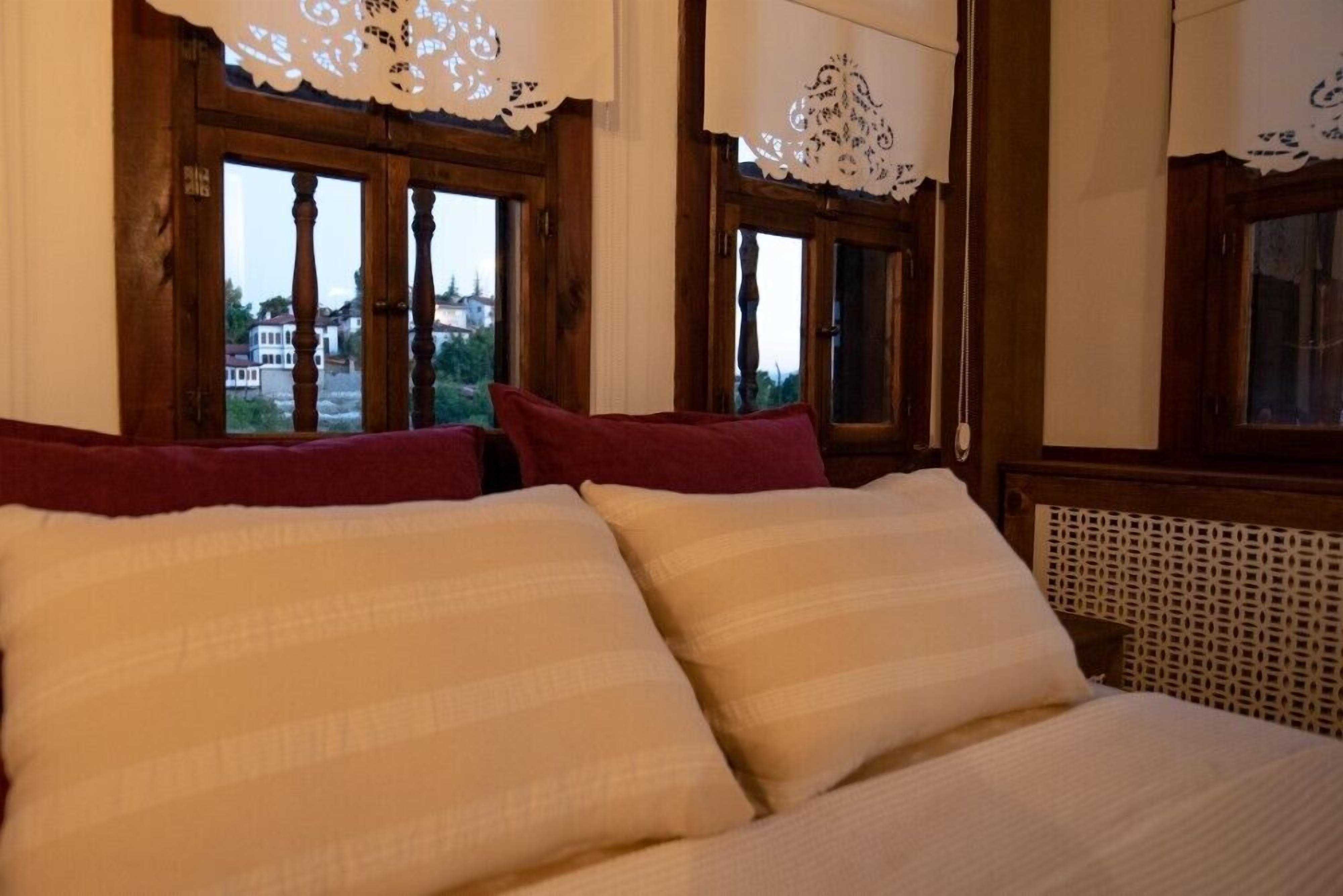 Una cama con almohadas blancas y rojas, marcos de ventana de madera y cortinas de encaje.
