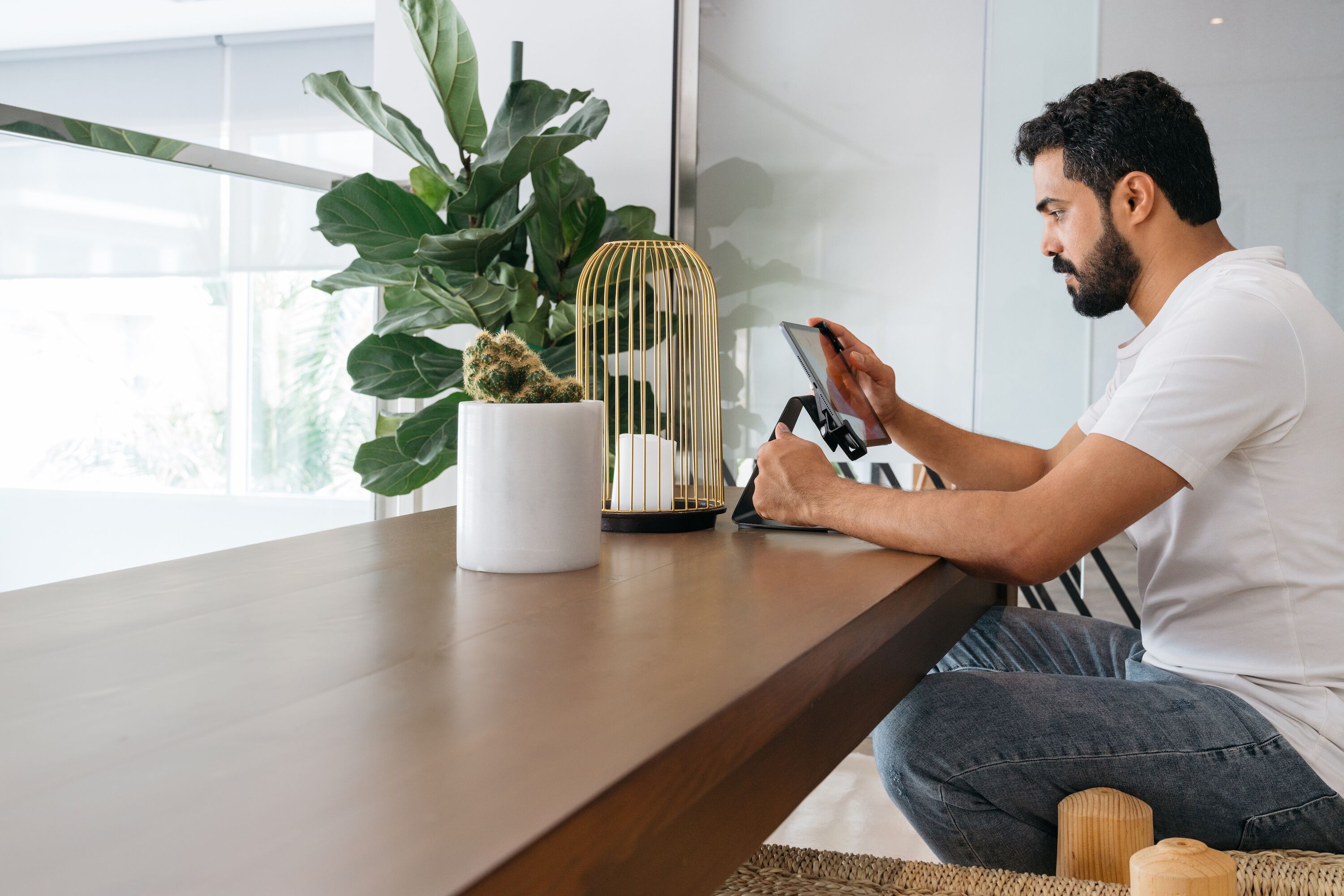 A person sitting at a wooden table with a smartphone, a white mug, a potted plant, and a gold cage with a cactus.