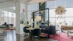 A modern office space with a glass partition, green chairs, a red rug, and a decorative chandelier. - Shada Hotel Shatea (Jeddah)