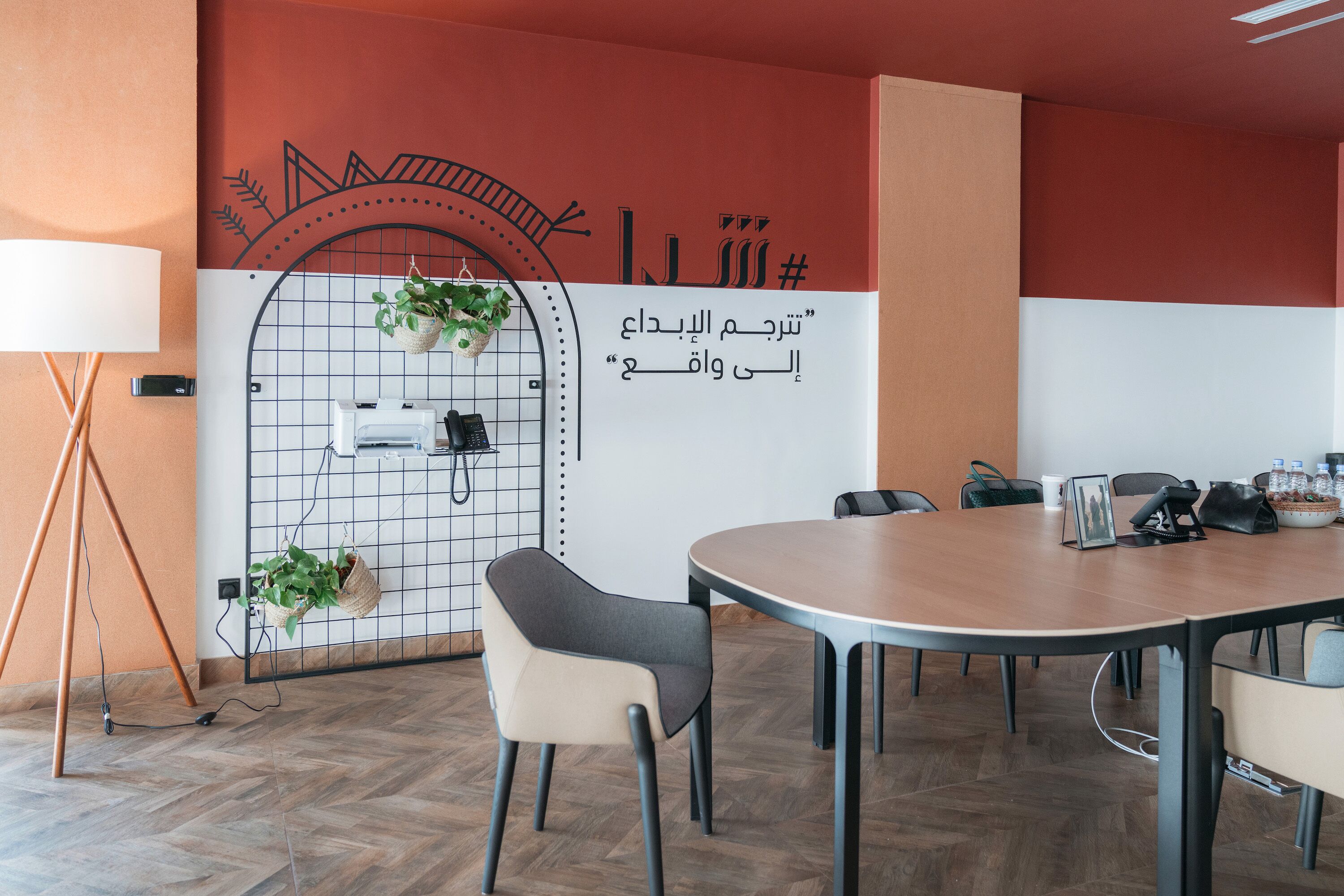 Une salle de conférence équipée d’une table ronde, de chaises, d’un tableau blanc sur lequel est écrit du texte en arabe, et de plantes en pot.