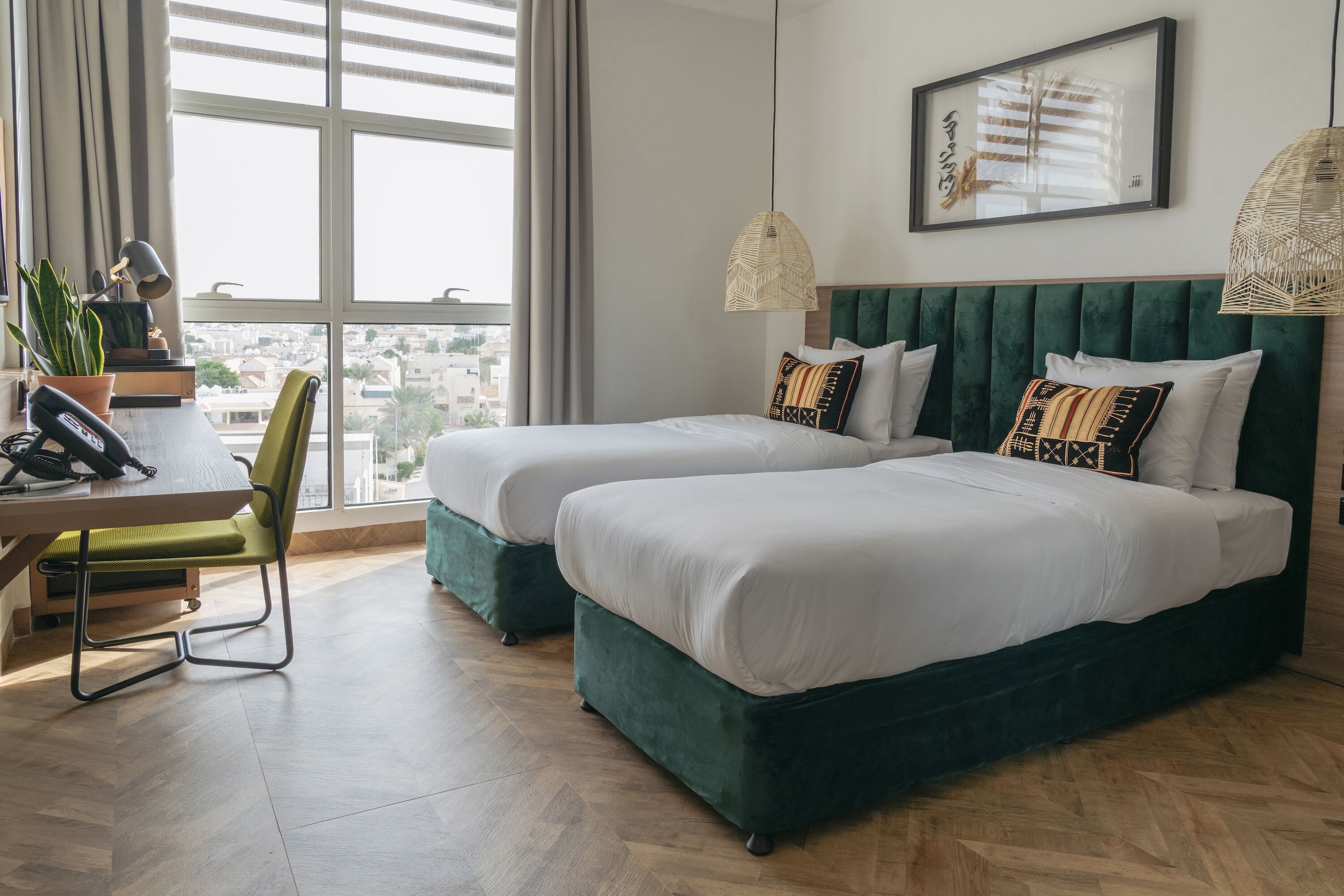 A hotel room with two beds, a desk with a chair, a phone, a plant, and a view of the city through large windows.