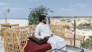 A man sitting on a wicker sofa with a red cushion, holding a smartphone, overlooking a cityscape with buildings and palm trees. - Shada Hotel Shatea (Jeddah)