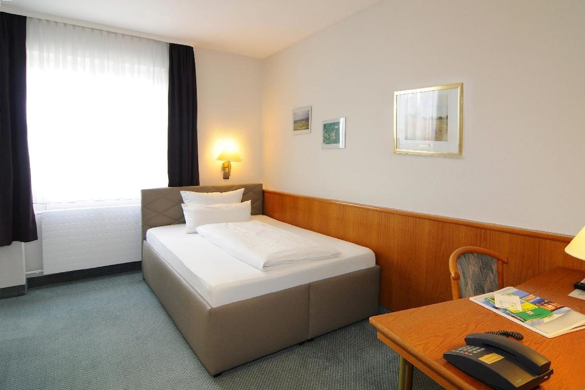 A hotel room with a bed, a desk with a telephone, a lamp, and a window with curtains.