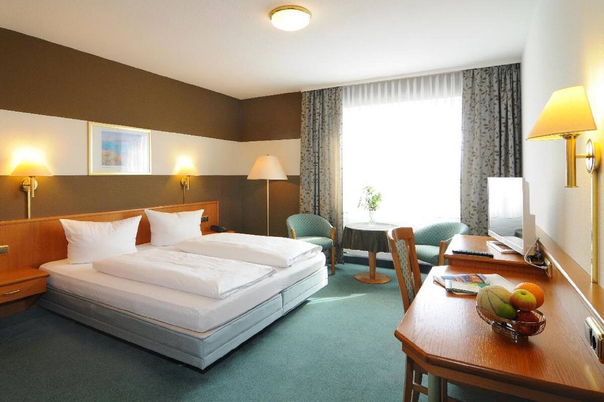 A hotel room with a double bed, bedside lamps, a desk with a chair, a small table with fruit, and a window with curtains.