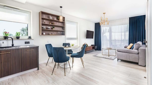 A modern living room with a grey sofa, a dining area with blue chairs, a kitchen with dark cabinets, and a large window with curtains. - Comfortable Apartment | Balcony and Parking in Garage (Kolobrzeg)