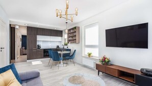 A modern living room with a sofa, coffee table, and TV. There is a dining area with chairs and a table. The kitchen is visible in the background. - Comfortable Apartment | Balcony and Parking in Garage (Kolobrzeg)