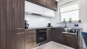 A modern kitchen with dark wood cabinets, white countertops, a built-in oven, and a window with blinds. - Comfortable Apartment | Balcony and Parking in Garage (Kolobrzeg)