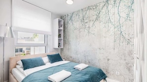 A modern bedroom with a large bed, a nightstand, a wall-mounted shelf, and a window with a view. - Comfortable Apartment | Balcony and Parking in Garage (Kolobrzeg)