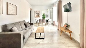 Una sala de estar moderna con un sofá, una mesa de centro y una televisión. En el fondo, hay un comedor con sillas y una mesa.