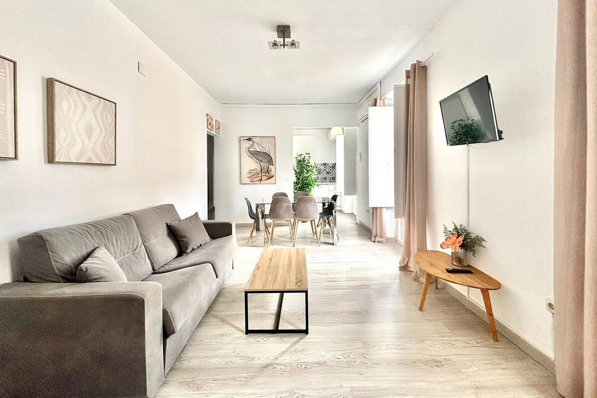 A modern living room with a sofa, coffee table, and TV. There is a dining area with chairs and a table in the background.