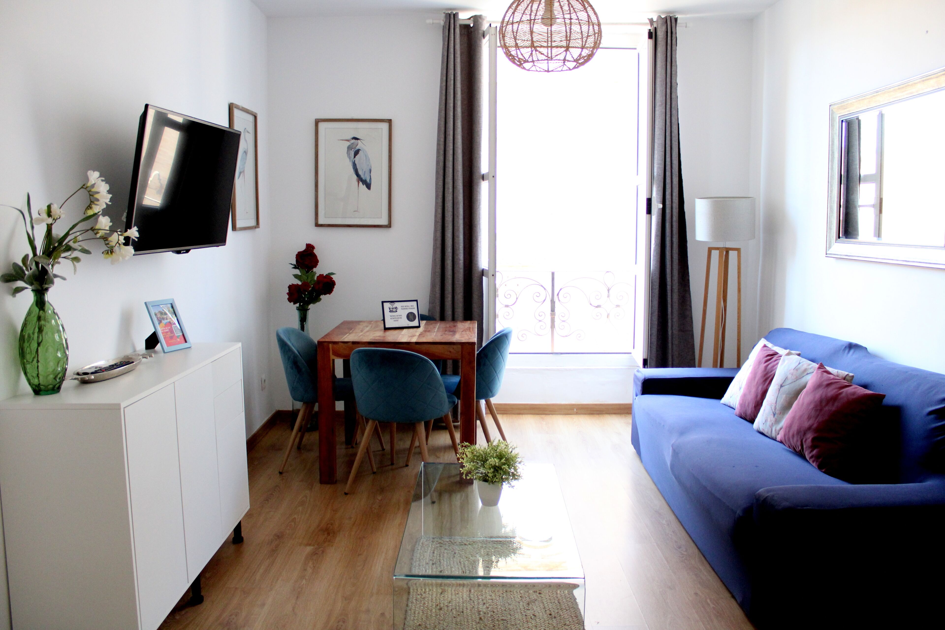 A modern living room with a sofa, a TV mounted on the wall, a dining table with chairs, and a small balcony with a view.