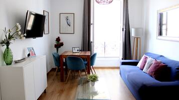 A modern living room with a sofa, a TV mounted on the wall, a dining table with chairs, and a small balcony with a view.