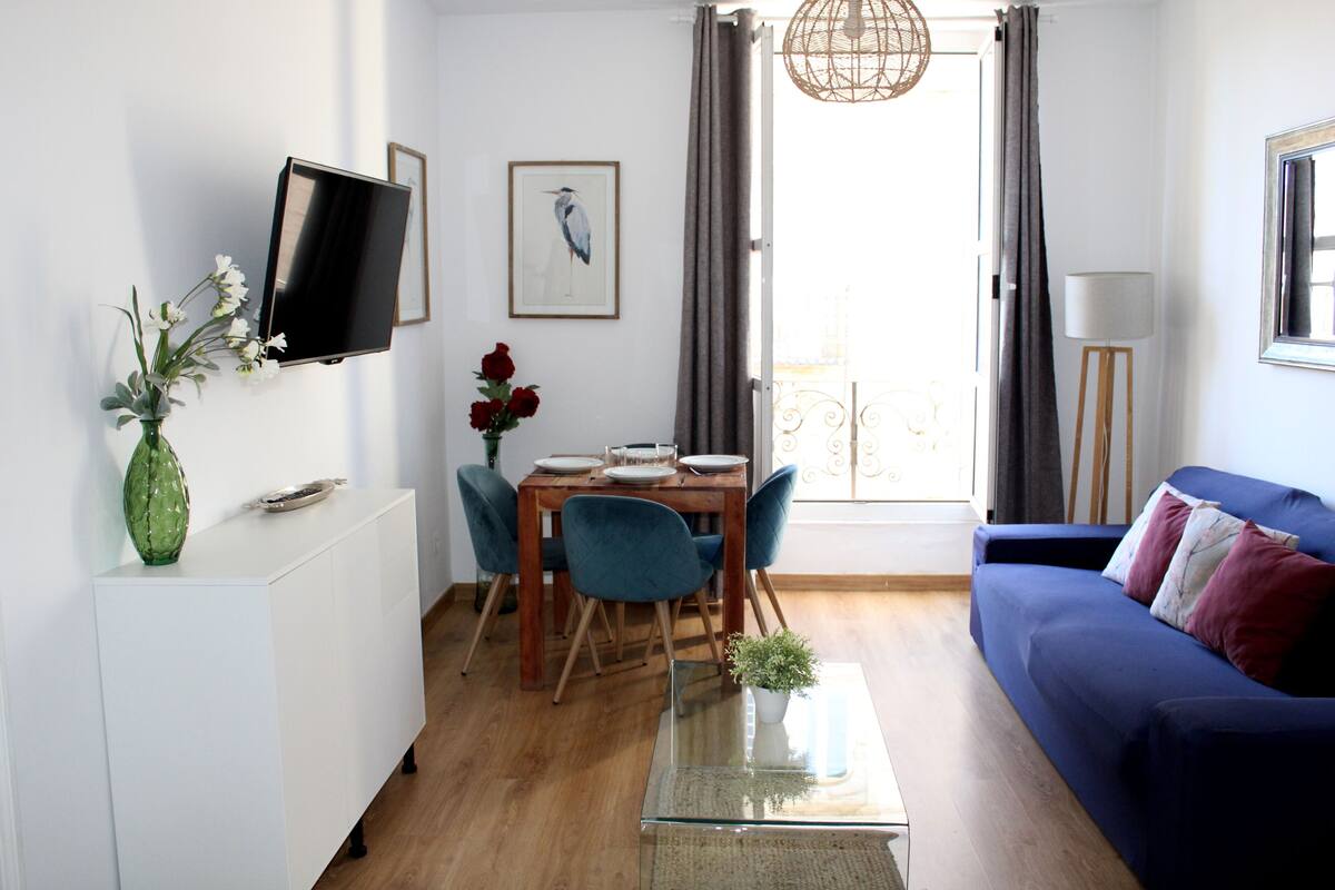 A living room with a blue sofa, a dining table with chairs, a TV mounted on the wall, and a window with curtains.