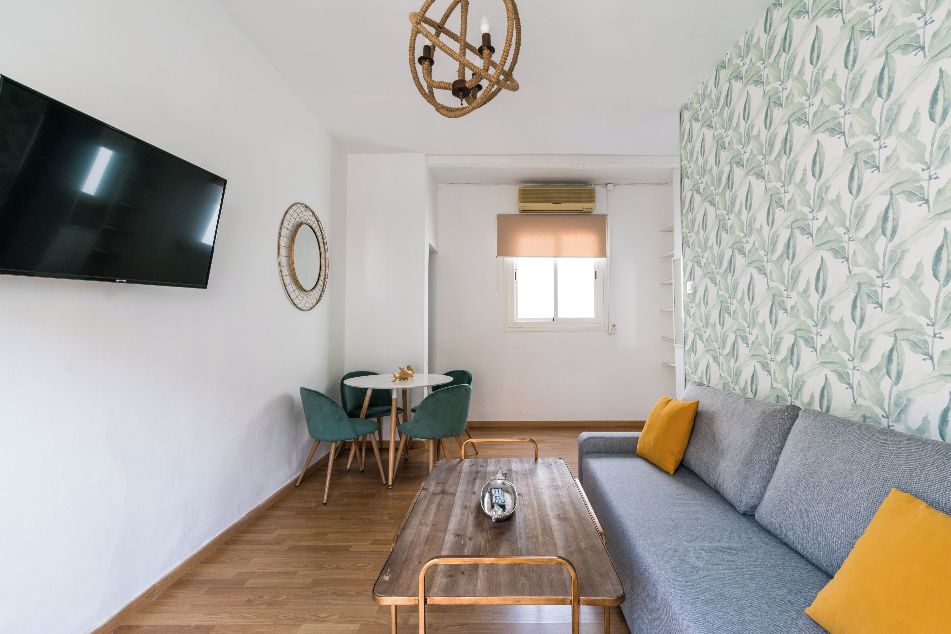Una sala de estar moderna con un televisor de pantalla plana, una mesa de centro de madera, un sofá gris con cojines amarillos y una zona de comedor con sillas verdes.