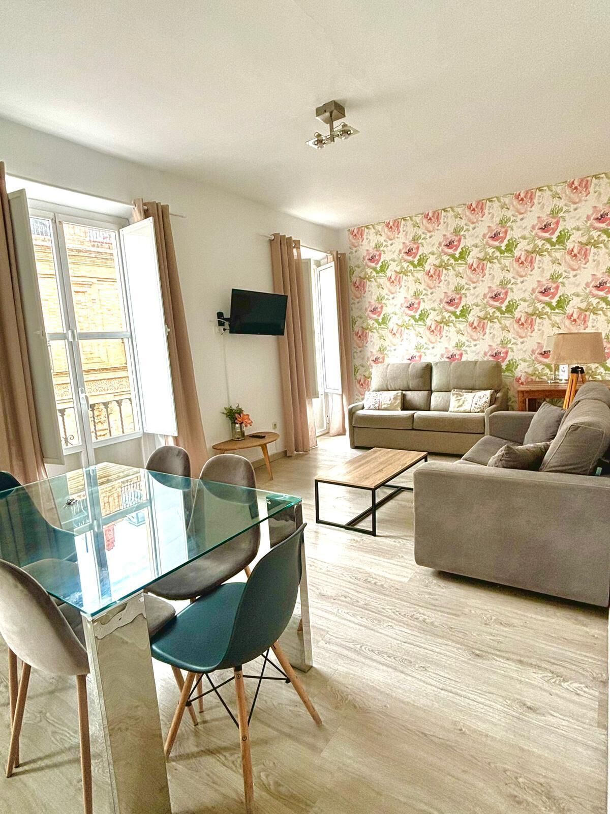 A living room with a sofa, a glass dining table with chairs, a TV mounted on the wall, and a floral wallpaper.