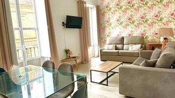 A living room with a sofa, a glass dining table with chairs, a TV mounted on the wall, and a floral wallpaper.