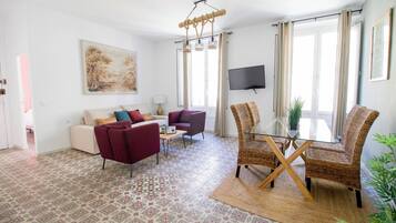 Un salon comprenant un canapé, un fauteuil, une table à manger et des chaises, un téléviseur fixé au mur et une grande fenêtre avec des rideaux.
