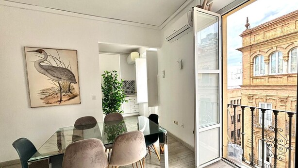 Un comedor moderno con una mesa y sillas de vidrio, una obra de arte enmarcada de un pájaro, una planta en maceta y vista a un edificio histórico desde un balcón.