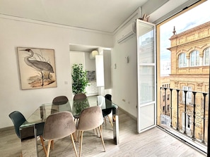 Un comedor moderno con una mesa y sillas de vidrio, una obra de arte enmarcada de un pájaro, una planta en maceta y vista a un edificio histórico desde un balcón.
