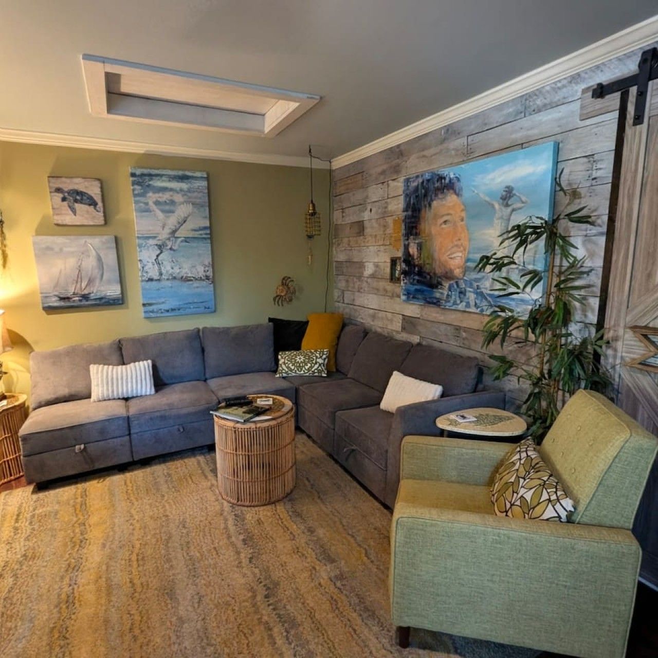 Ein Wohnzimmer mit einem Ecksofa, einem runden Couchtisch und einem grünen Sessel. An den Wänden befinden sich Wandbilder und ein großes Gemälde auf Leinwand.