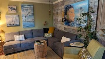 Ein Wohnzimmer mit einem Ecksofa, einem runden Couchtisch und einem grünen Sessel. An den Wänden befinden sich Wandbilder und ein großes Gemälde auf Leinwand.
