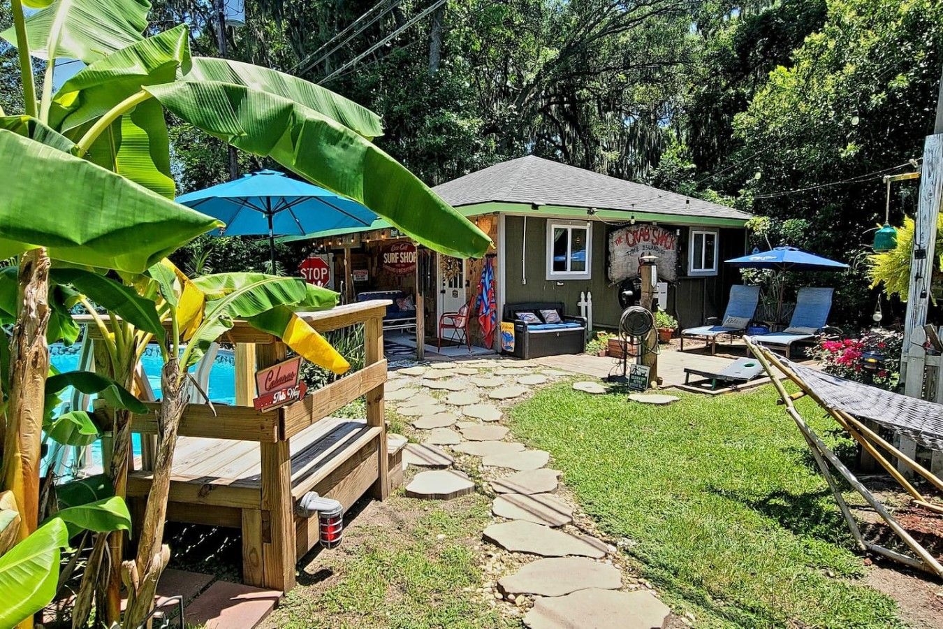 A backyard with a wooden deck, a pool, and a small building with a sign that reads 'The Good Stuff Surf Shop.'
