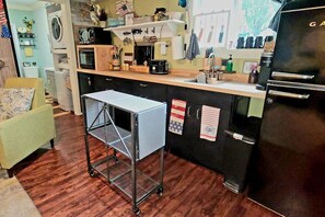 Eine Küche mit Mikrowelle, Kaffeemaschine und Kühlschrank. Ein kleiner Tisch mit Metallgestell und Rollen steht bereit.