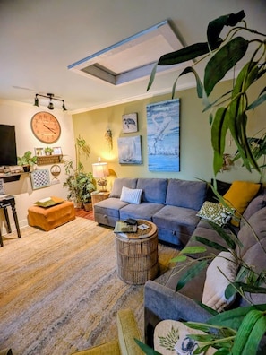 A living room with a sofa, ottoman, and a round coffee table. There are wall-mounted paintings and a clock.