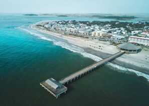An aerial view of a coastal area with a long pier extending into the water, a beach, and a cluster of buildings.