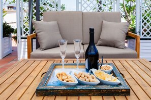Ett träbord med ett bricka som innehåller snacks och en vinflaska, placerat på en uteplats med en soffa och en spaljé som bakgrund.
