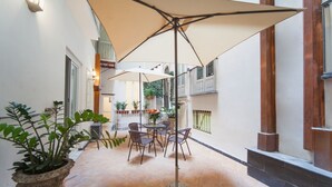 Een binnenplaats met een tafel en stoelen onder een grote parasol, omringd door potplanten en een betegelde vloer.