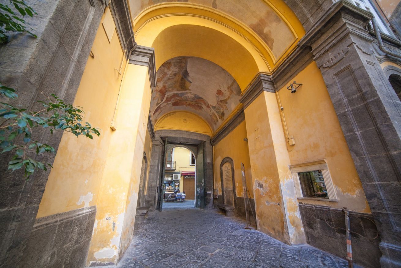 An arched corridor with stone flooring and yellow walls, leading to a doorway.