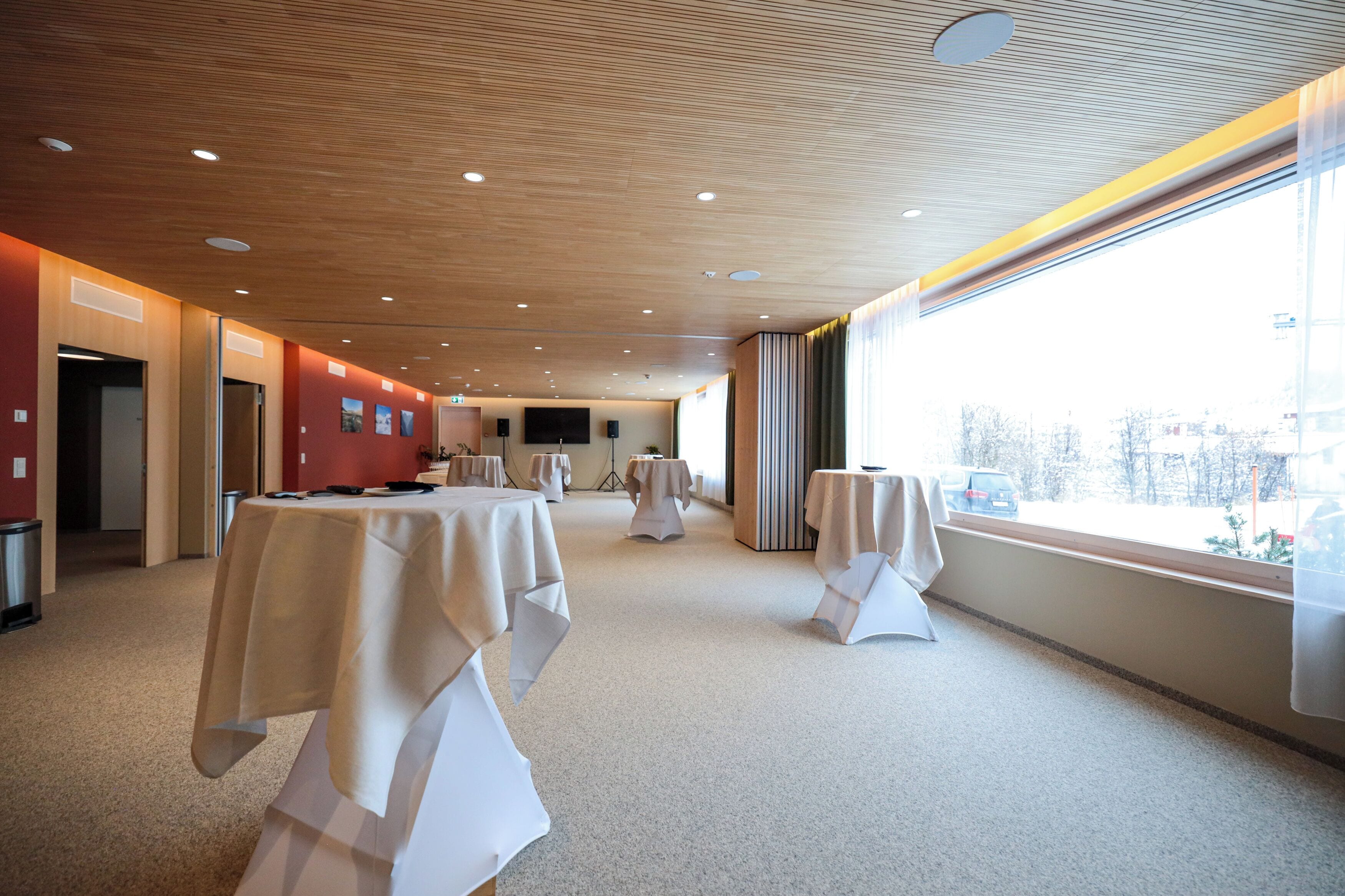 Un salón de conferencias con mesas dispuestas para una reunión, un ventanal con vistas a la ciudad y un techo de madera con iluminación empotrada.