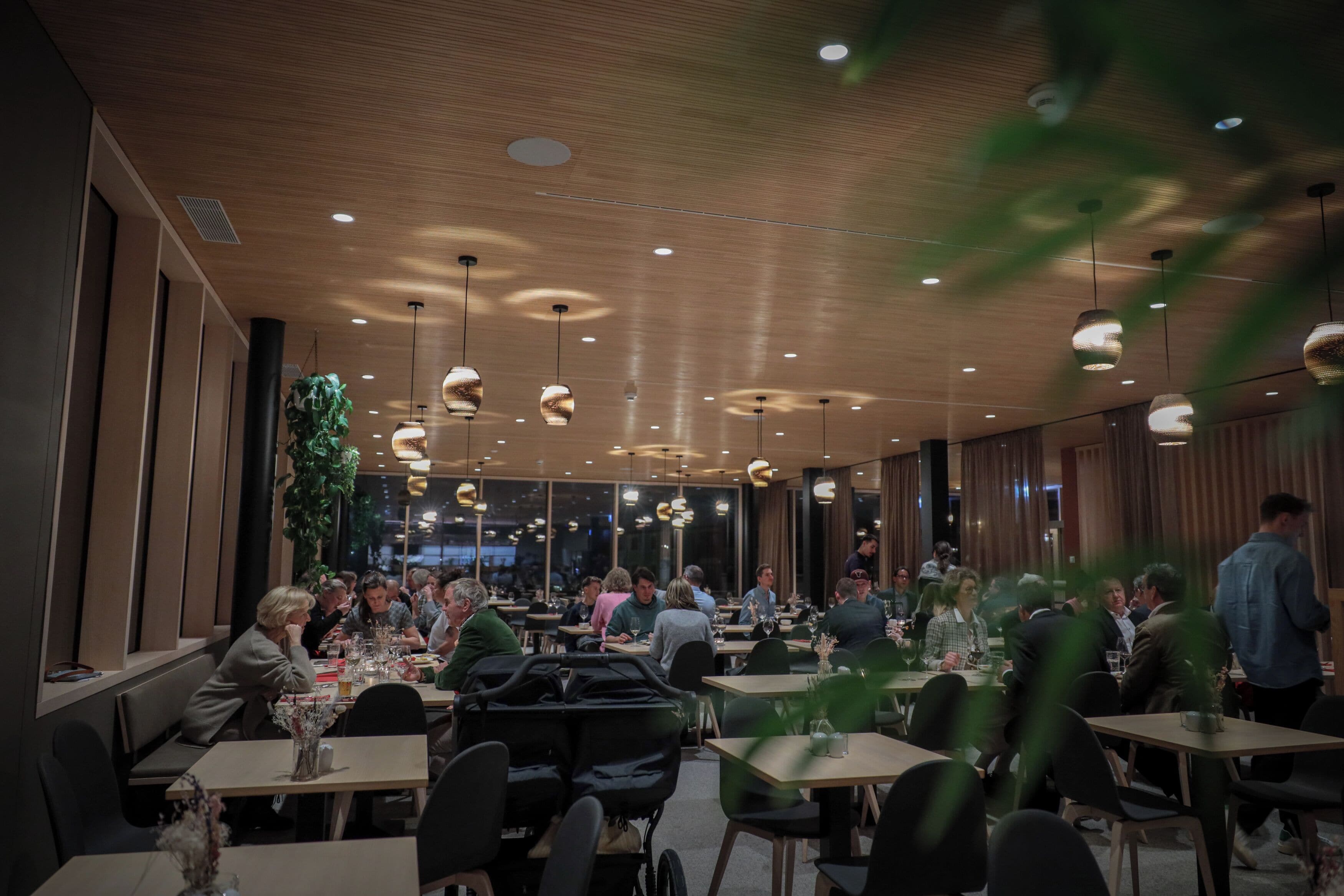 Restaurante com teto de madeira e iluminação pendente, mesas com clientes e grandes janelas.