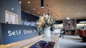 A reception desk with a 'Self Check In' sign, a tablet, and a floral arrangement. - AVES Hotel Savognin (Surses)