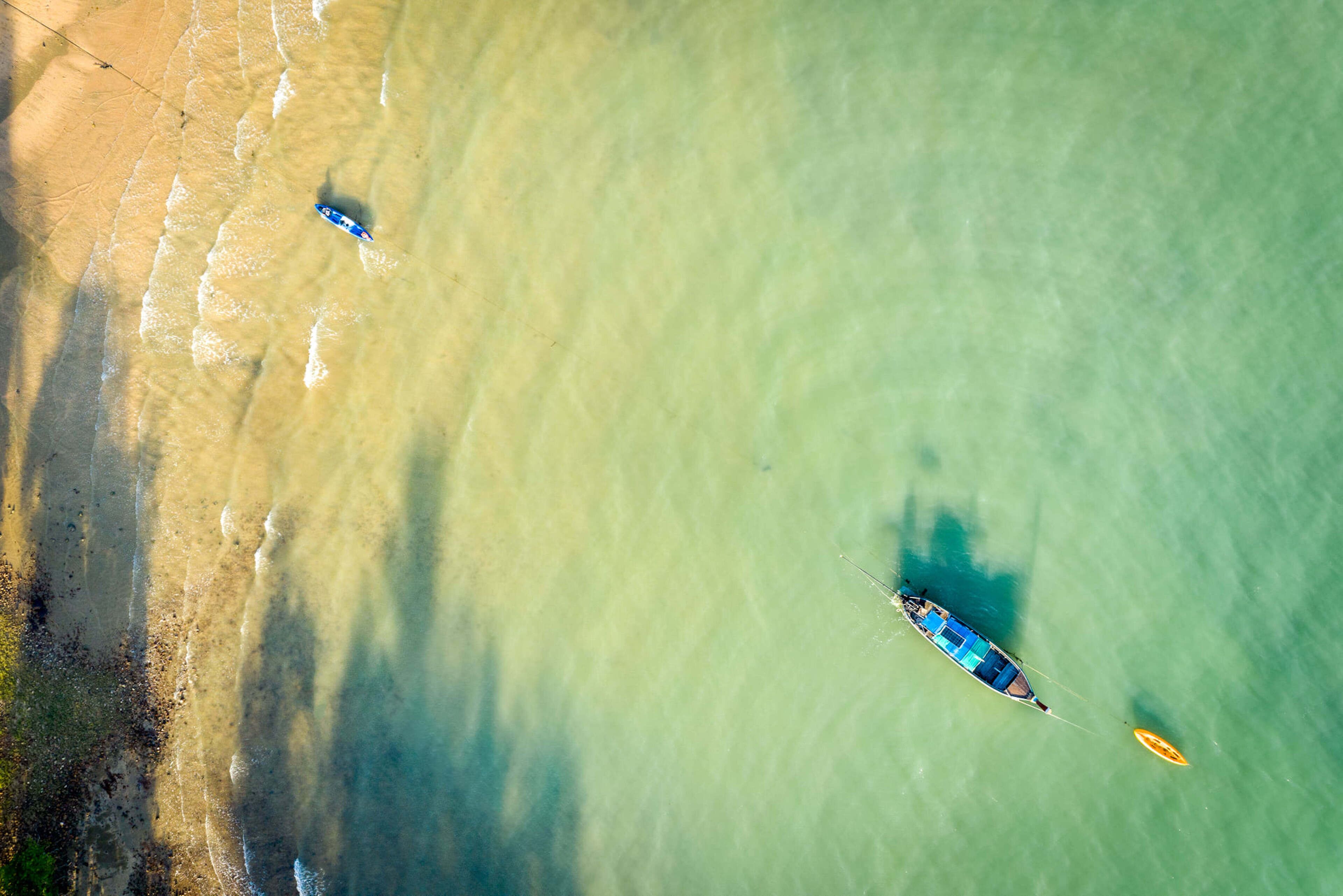 An aerial view of a beach with clear water, a boat, and a surfboard.