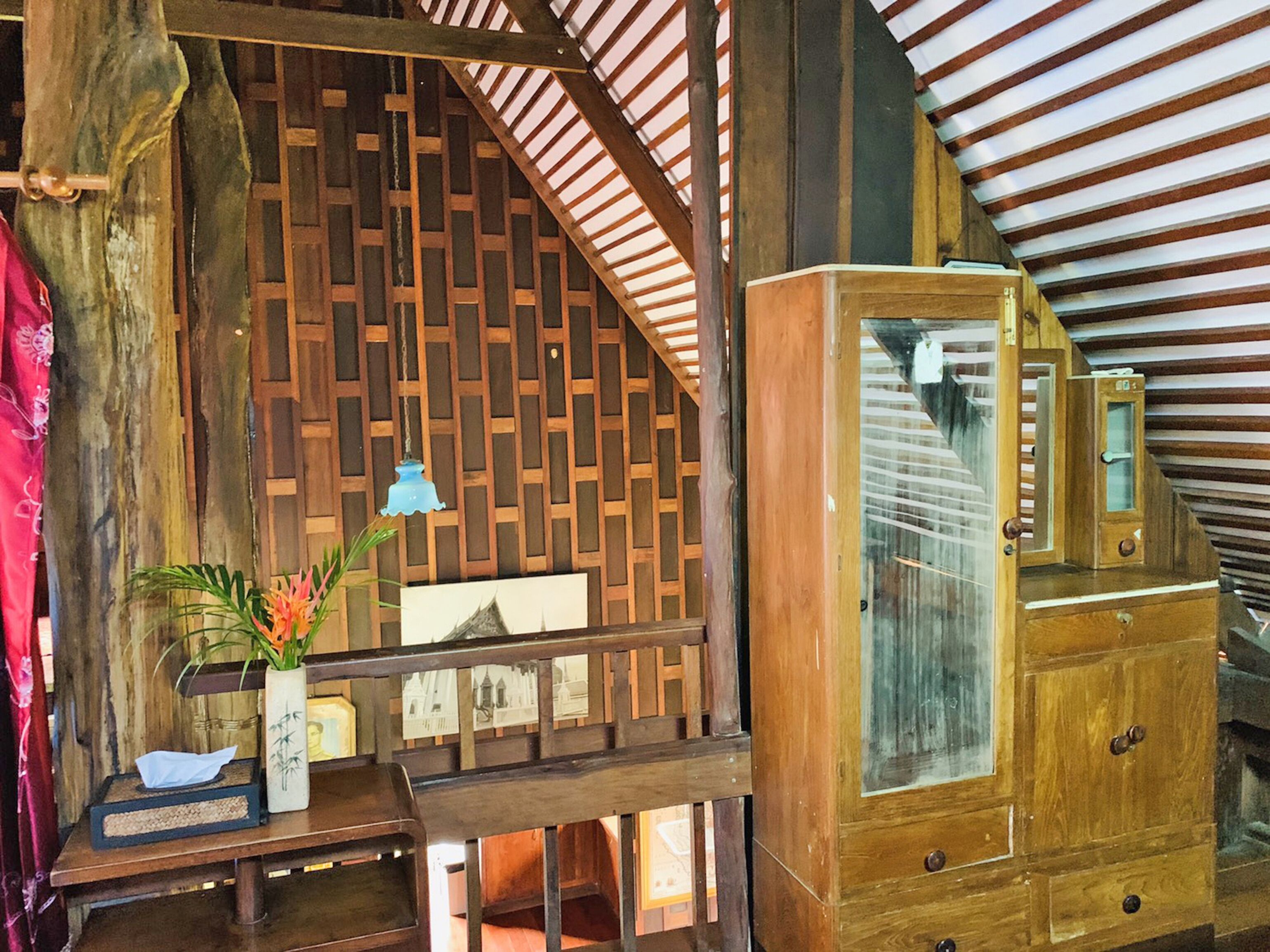 A rustic wooden room with a sloped ceiling, a wooden cabinet with glass doors, and a small table with a lamp and a plant.