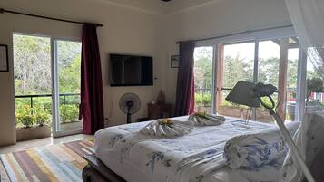A bedroom with a large bed, a flat-screen TV, a ceiling fan, and a view of greenery through the windows.
