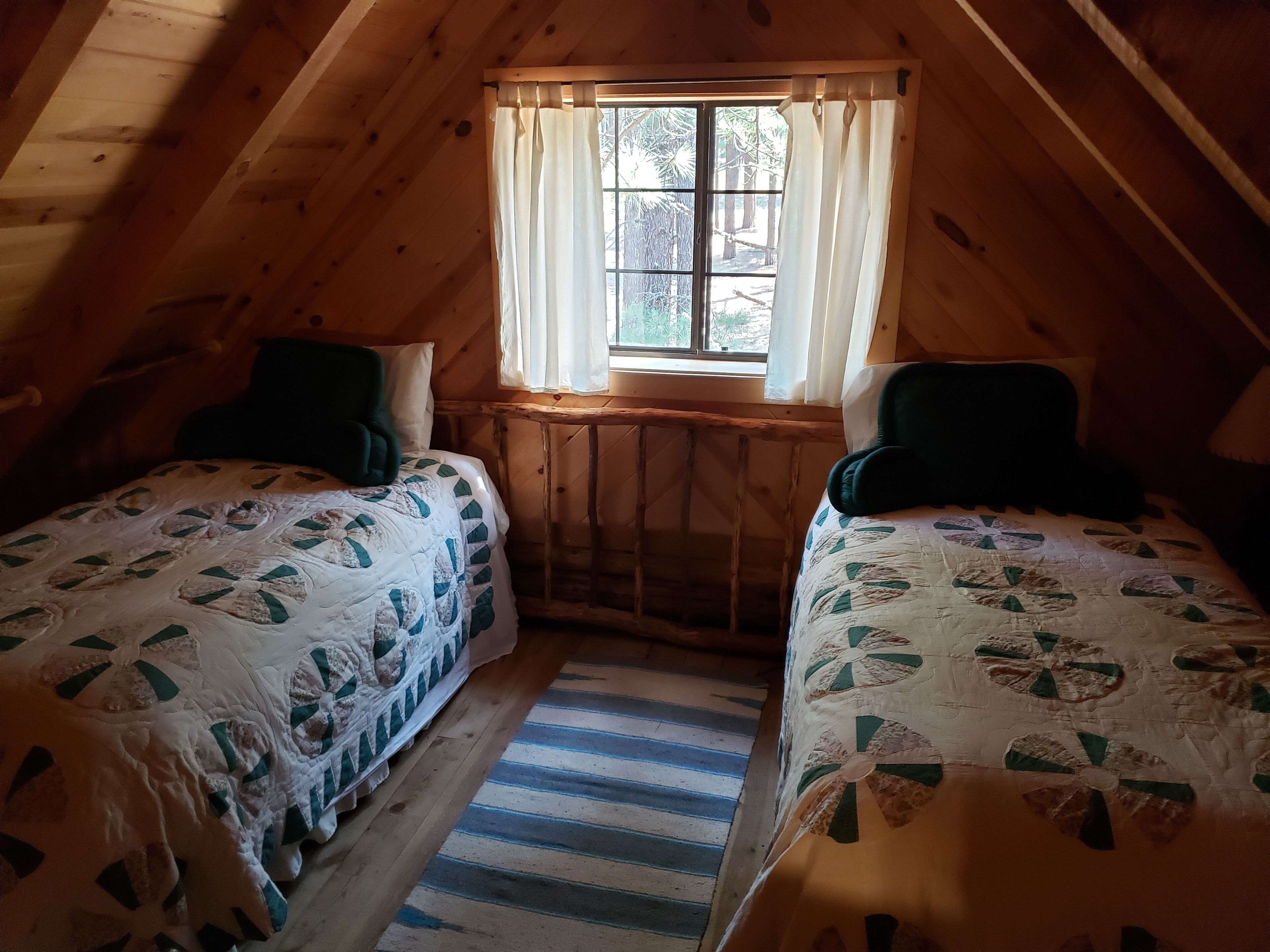 A cozy attic bedroom with two beds, a window with curtains, and a wooden ceiling.