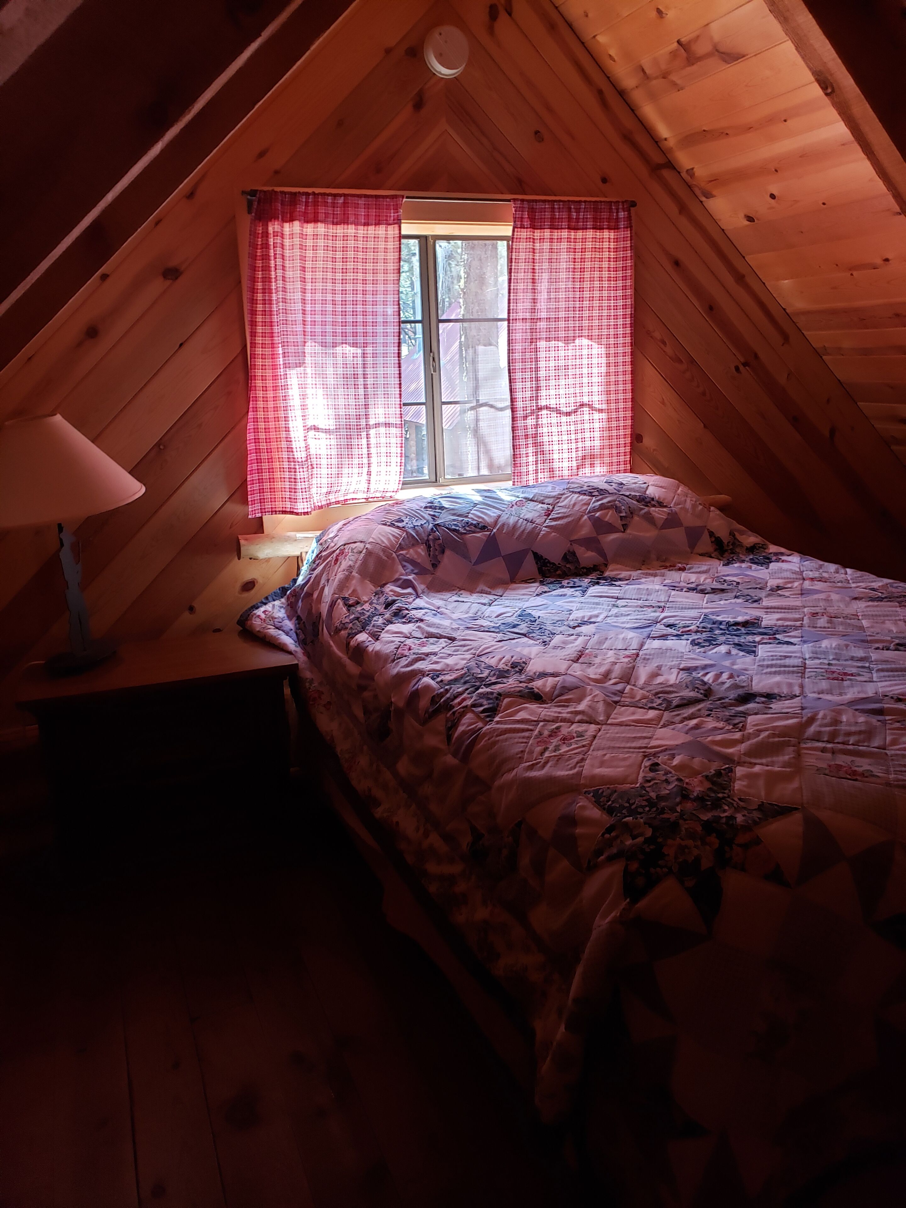 A cozy attic bedroom with a wooden sloped ceiling, a bed with a patterned quilt, a nightstand with a lamp, and a window with red curtains.
