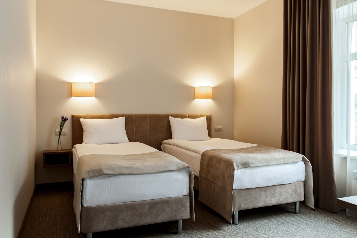 Une chambre d’hôtel avec deux lits, une table de chevet, une lampe et une fenêtre avec des rideaux.
