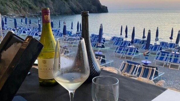 A table with a bottle of wine, a wine glass, and a menu on a terrace overlooking a beach with blue chairs and umbrellas. - Elegant apartment with view of the sea (Piombino)