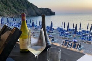 Een tafel met een fles wijn, een wijnglas en een menukaart op een terras met uitzicht op een strand, met blauwe stoelen en parasols.