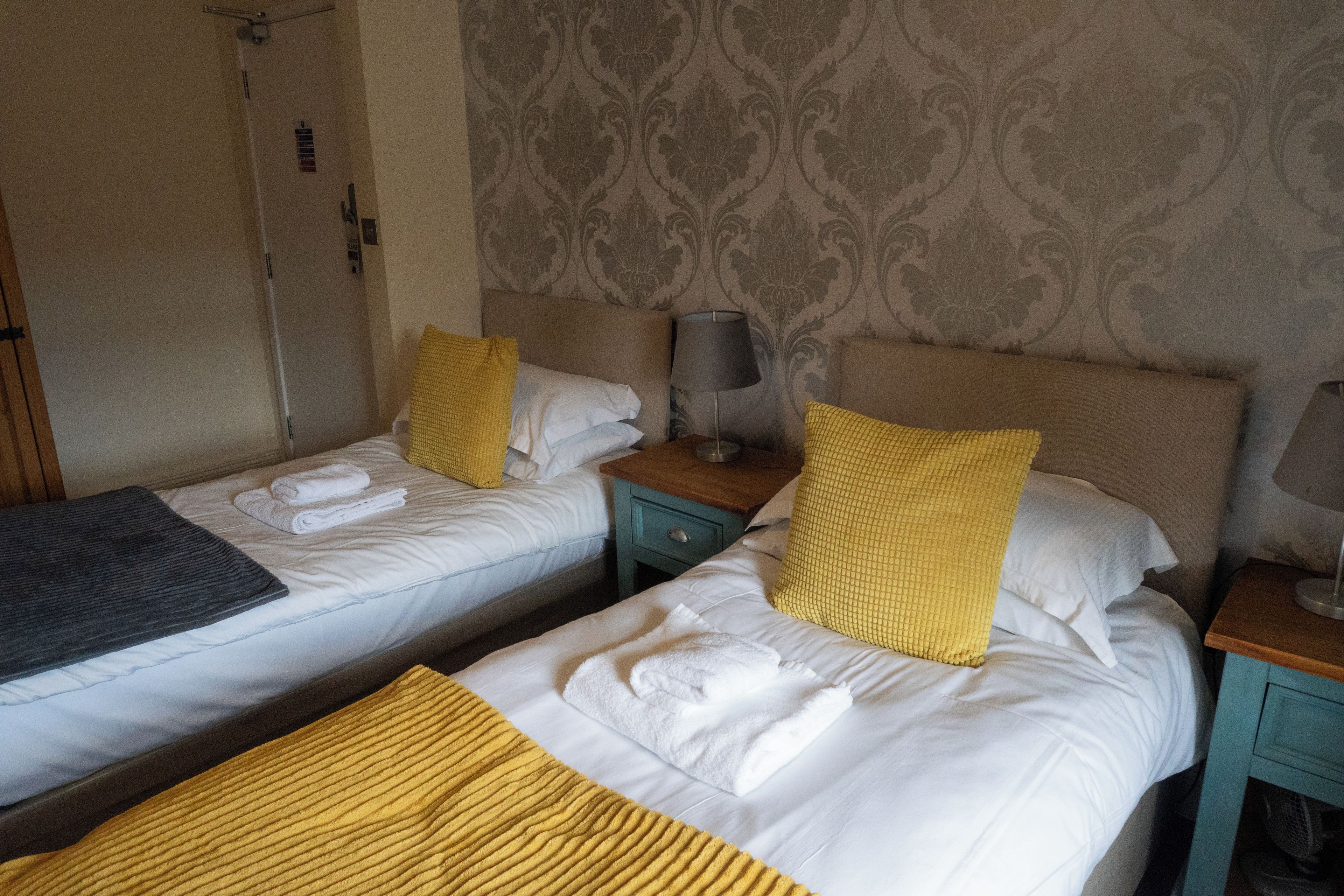 Quarto de hotel com duas camas de solteiro, cada uma com roupa de cama branca, um cobertor amarelo e almofadas combinando. Há uma mesa de cabeceira com uma luminária entre as camas.