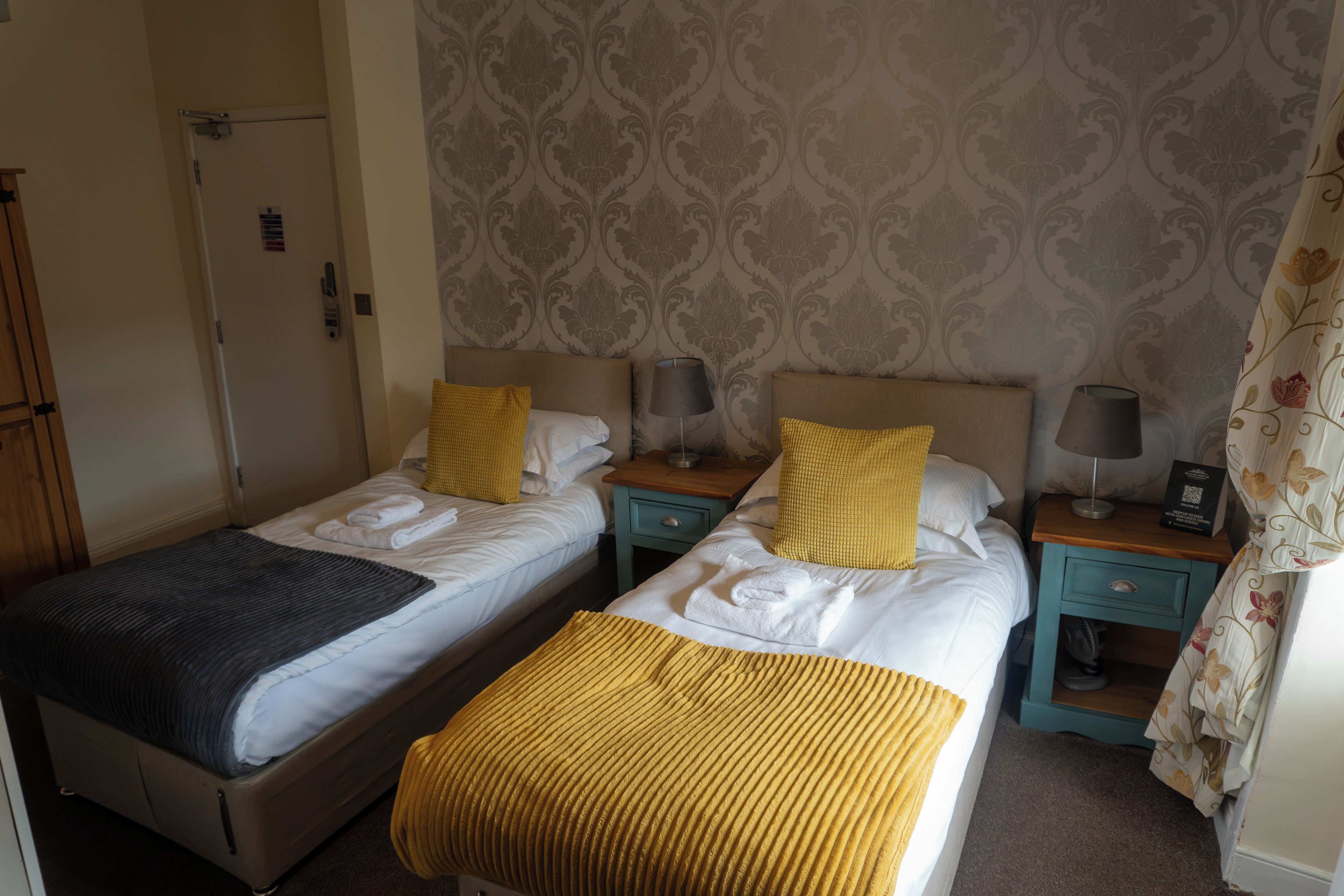 A hotel room with two beds, each with a pillow and a lamp. There is a door and a wall with a patterned design.